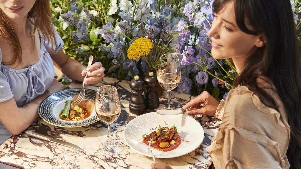Two women eat dinner outside with a backdrop of flowers