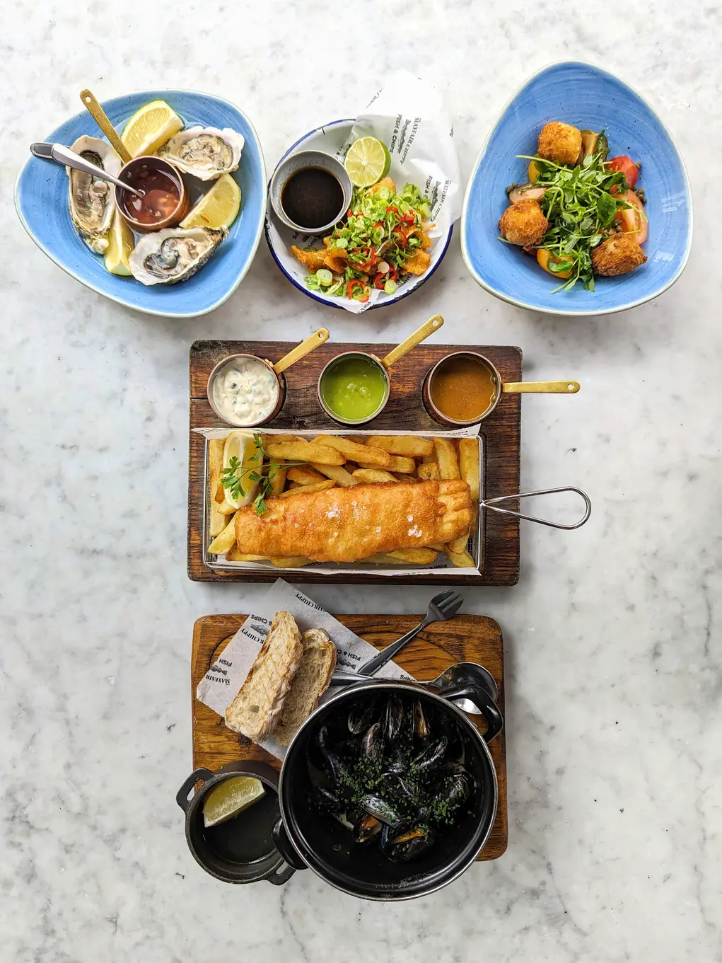 A table filled with five plates of food including fish and chips and mussels